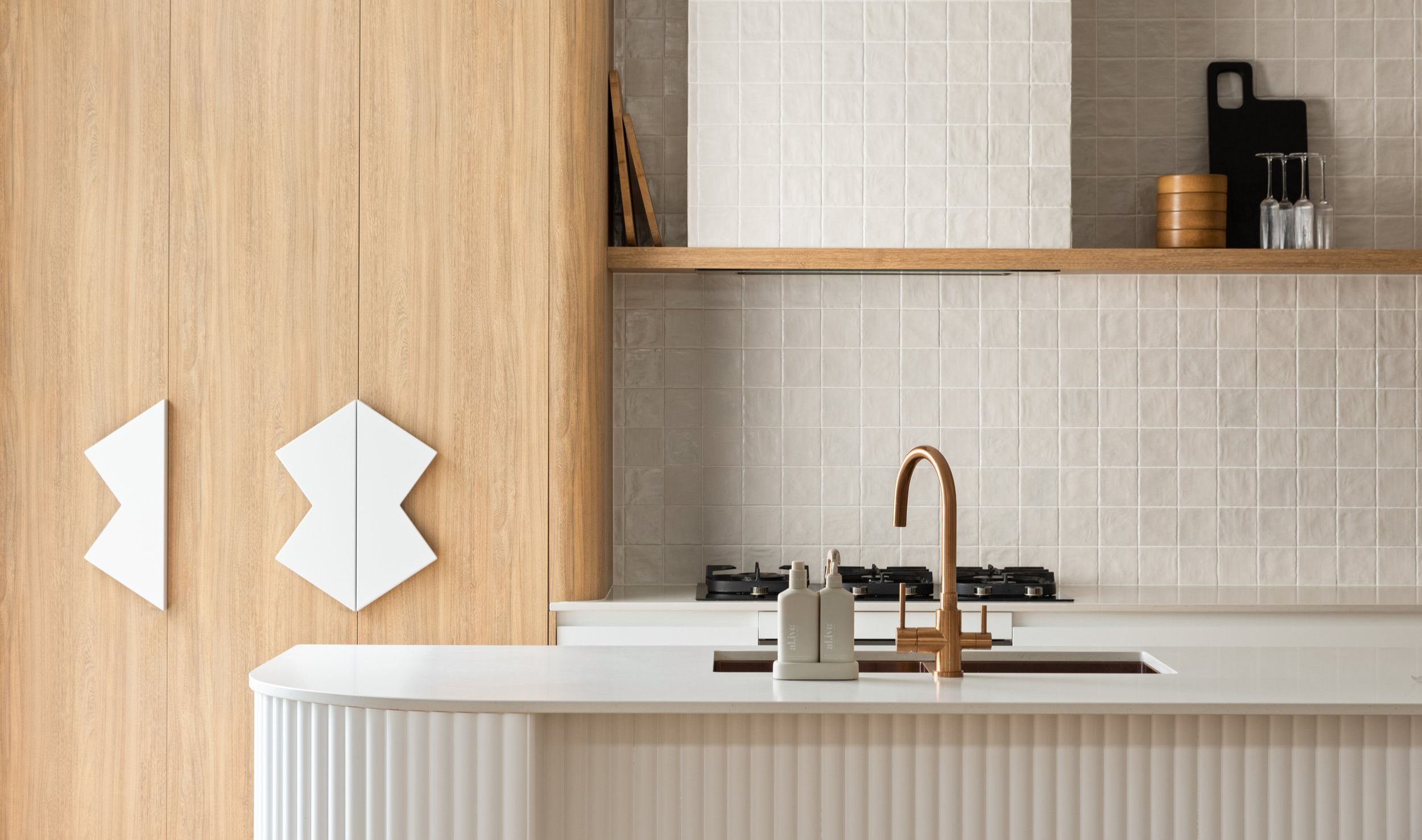 Modern kitchen with a white counter, copper faucet, beige tiled backsplash, wooden cabinets, and decorative items.