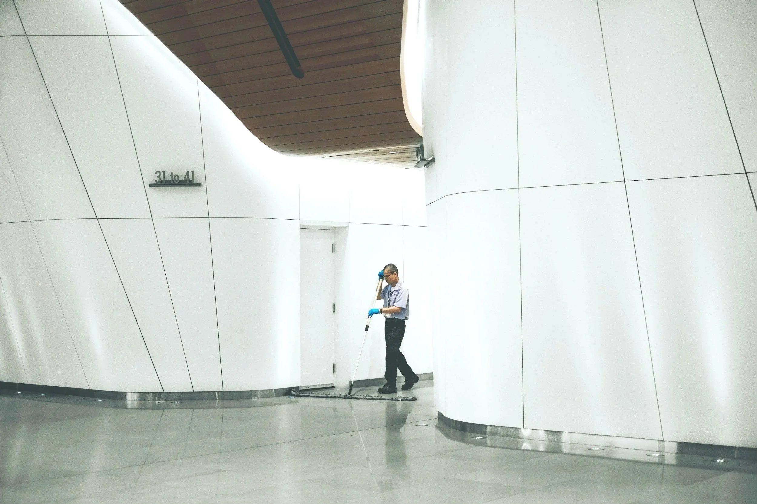 A man cleaning the floor in a modern, white-walled building with curved walls and a wooden ceiling.