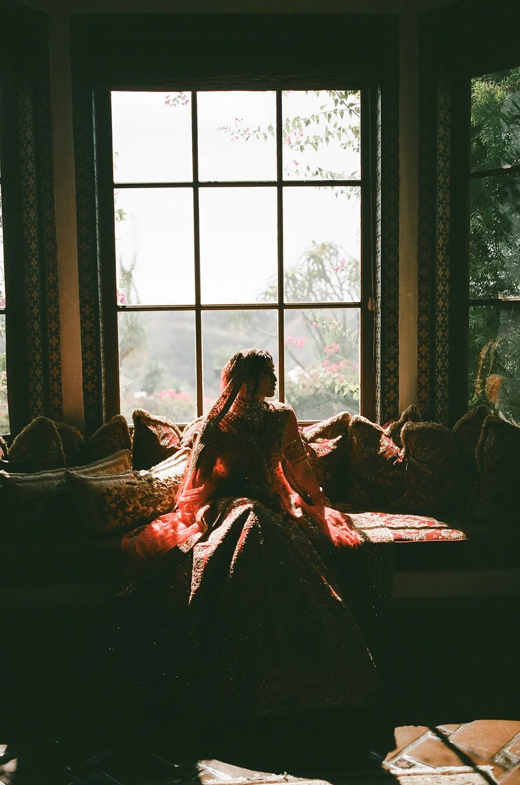 A woman wearing a traditional dress sitting on a cushioned window seat inside a room, backlit by sunlight coming through large window panes, with plants and trees visible outside.