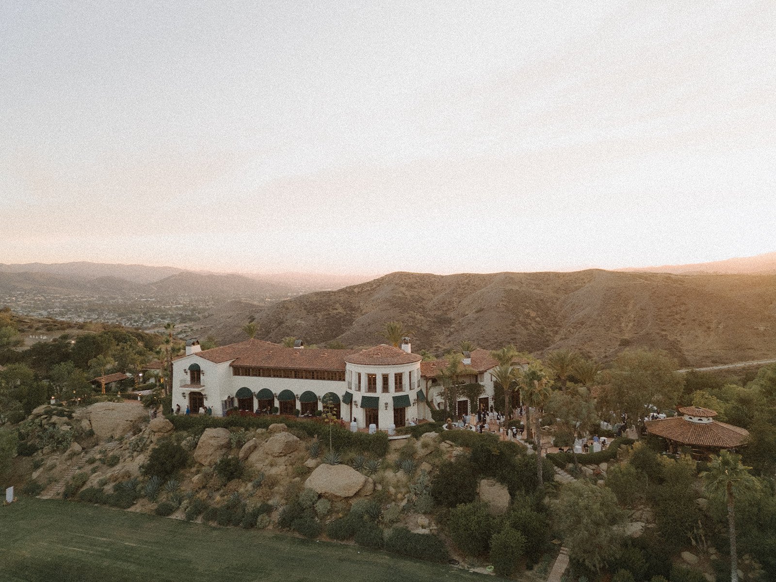 A large estate with white walls and a red-tiled roof, surrounded by trees and rocks, set against rolling hills at sunset.