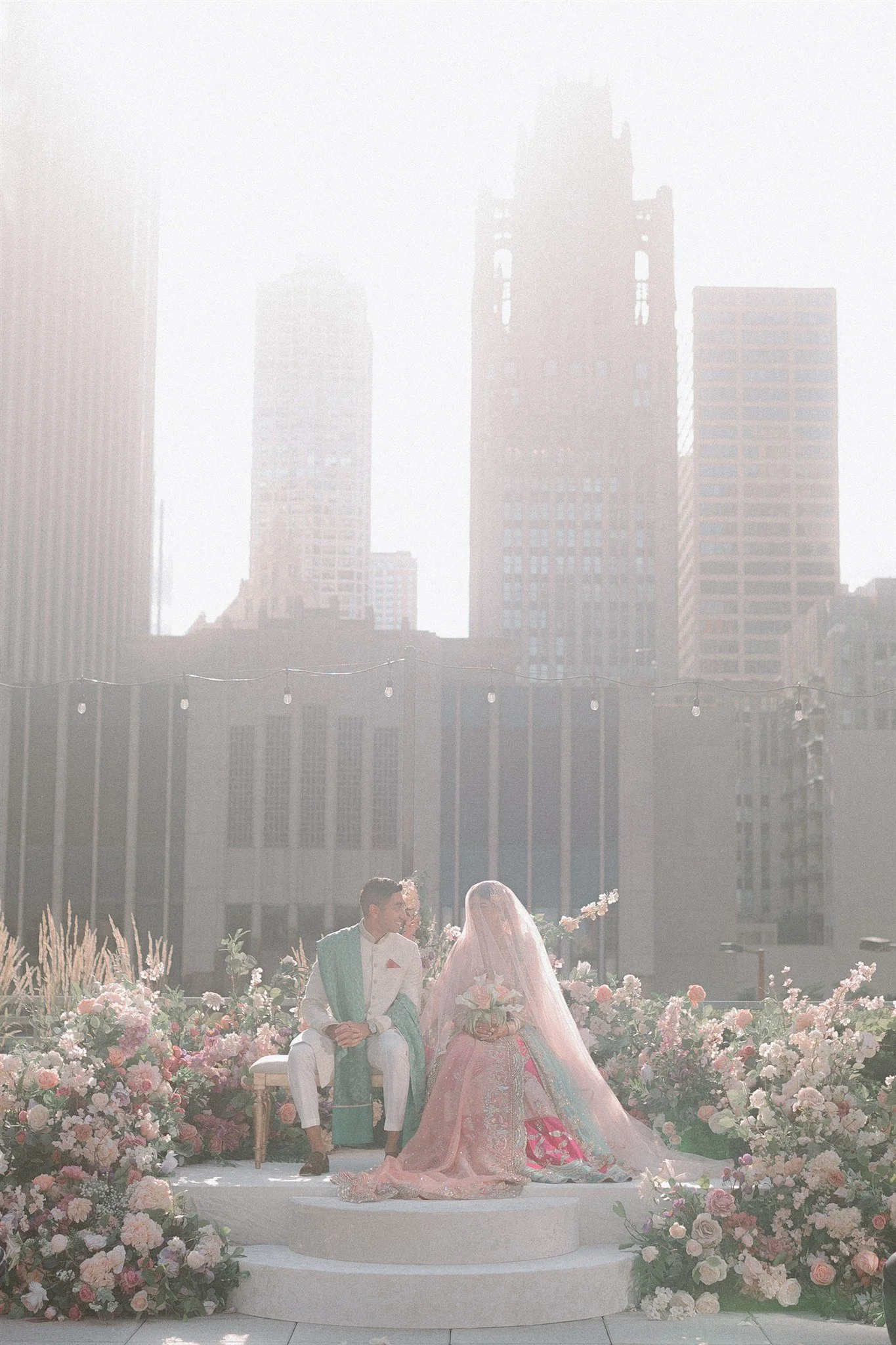 A wedding ceremony on a rooftop with a city skyline in the background, featuring a bride in a pink dress and veil, and a groom in a white suit with a turquoise sash.