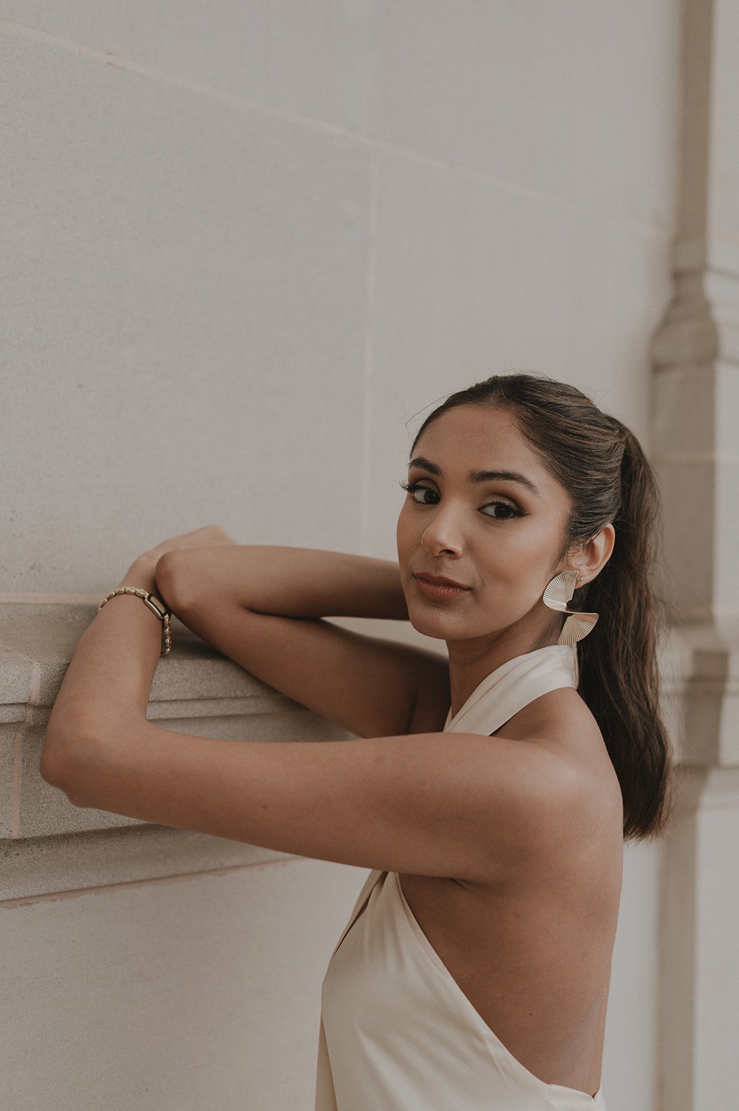 A young woman with dark hair in a ponytail, wearing a sleeveless cream-colored dress and large earrings, posing against a beige brick wall.