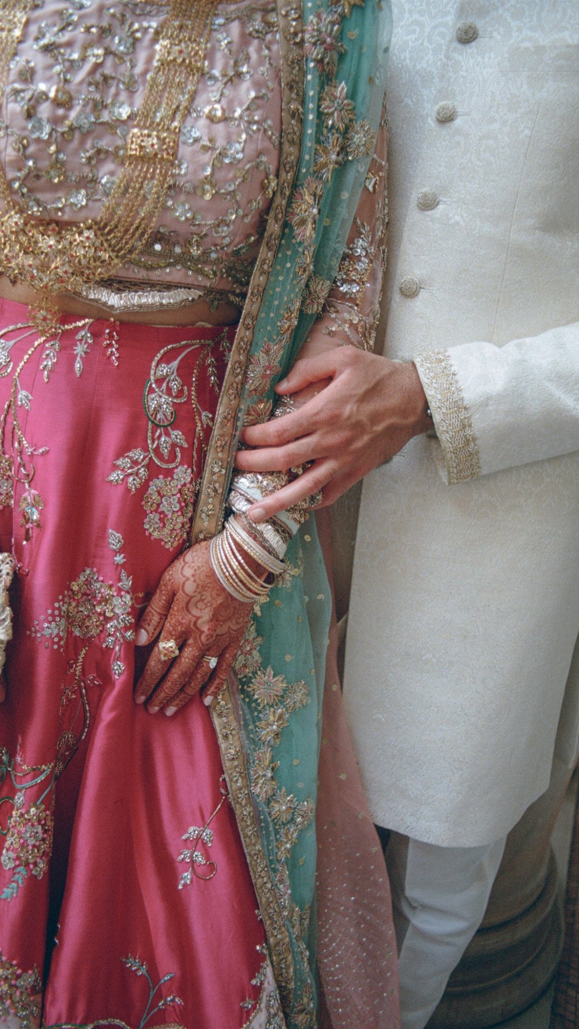 A bride in a pink and gold embroidered traditional wedding dress, adorned with jewelry, holding her hand, which is decorated with henna and bangles, while a person in white traditional attire gently touches her dress.