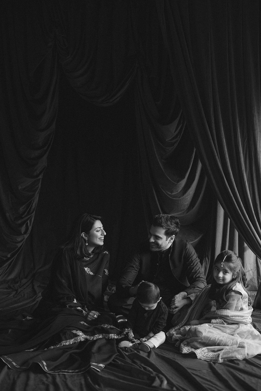 A family of four sitting on a bed with dark curtains in the background, smiling and interacting.