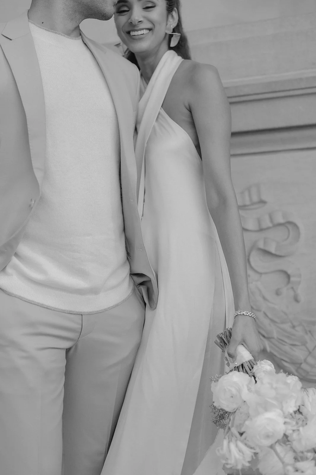 A black and white photo of a smiling woman in a wedding dress holding a bouquet, standing beside a man in light-colored suit pants and a light shirt. They are close together, with part of the man's face visible.
