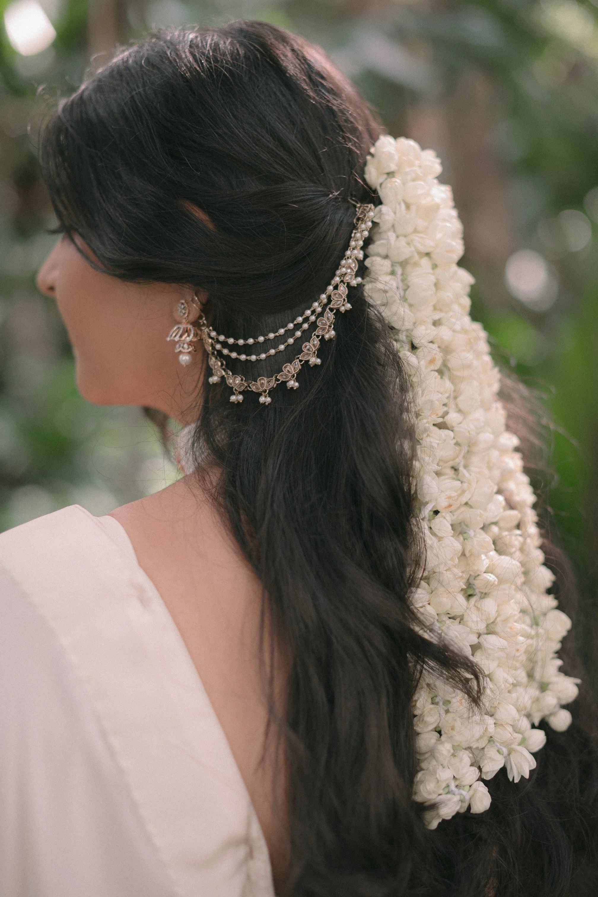 A woman with long black hair styled with silver jewelry and adorned with a large floral garland of white flowers, likely at a wedding or ceremonial event.