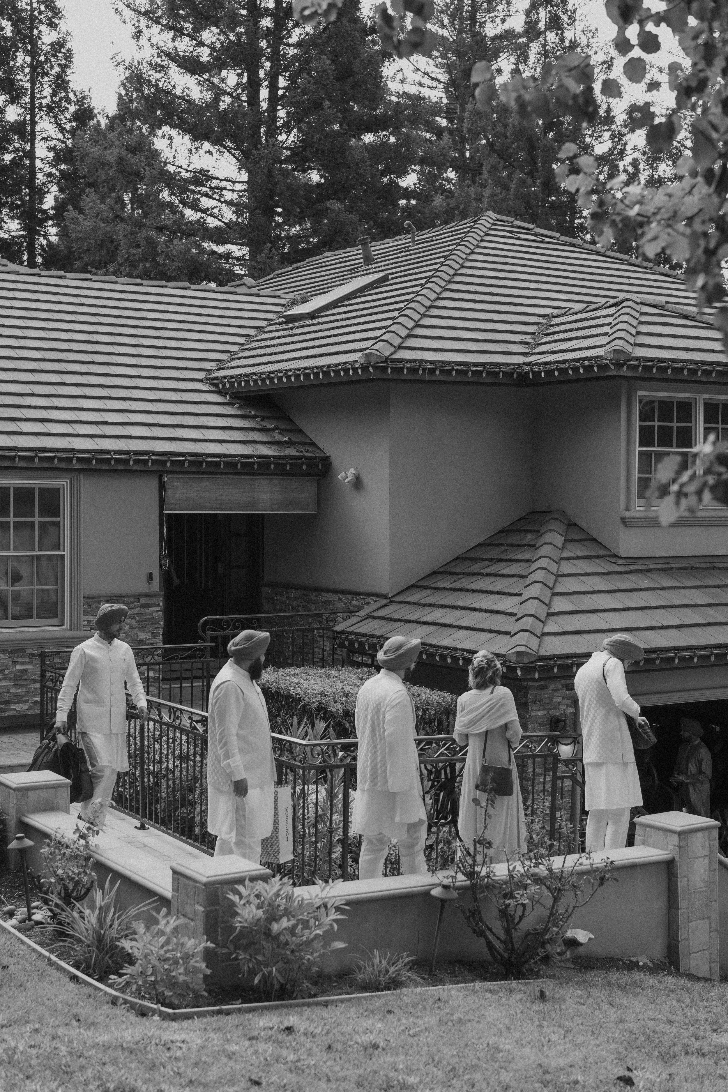 People waiting in line outside a house with a tiled roof, in a residential area with trees in the background.