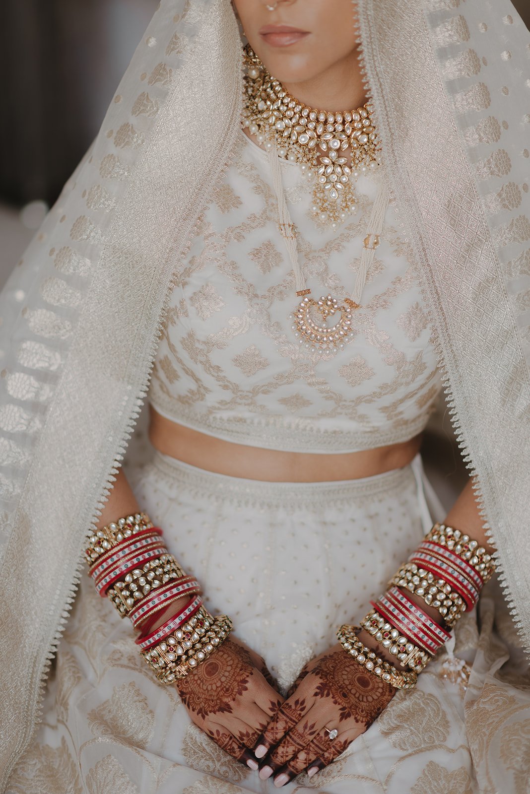 Close-up of a bride wearing traditional Indian attire and jewelry, including a white and gold embroidered blouse, multiple pearl and gold necklaces, and red and gold bangles. She has henna designs on her hands.