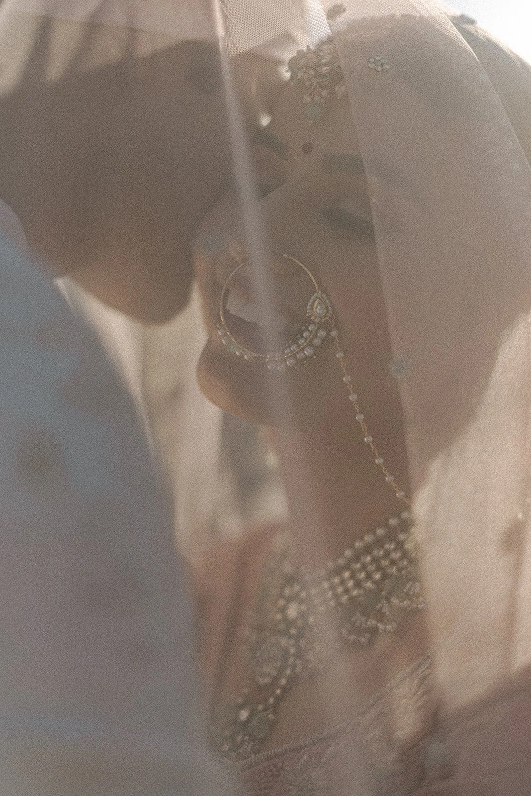 Close-up of a woman wearing jewelry, including earrings, a necklace, and a ring, with sunlight and a sheer veil fabric.