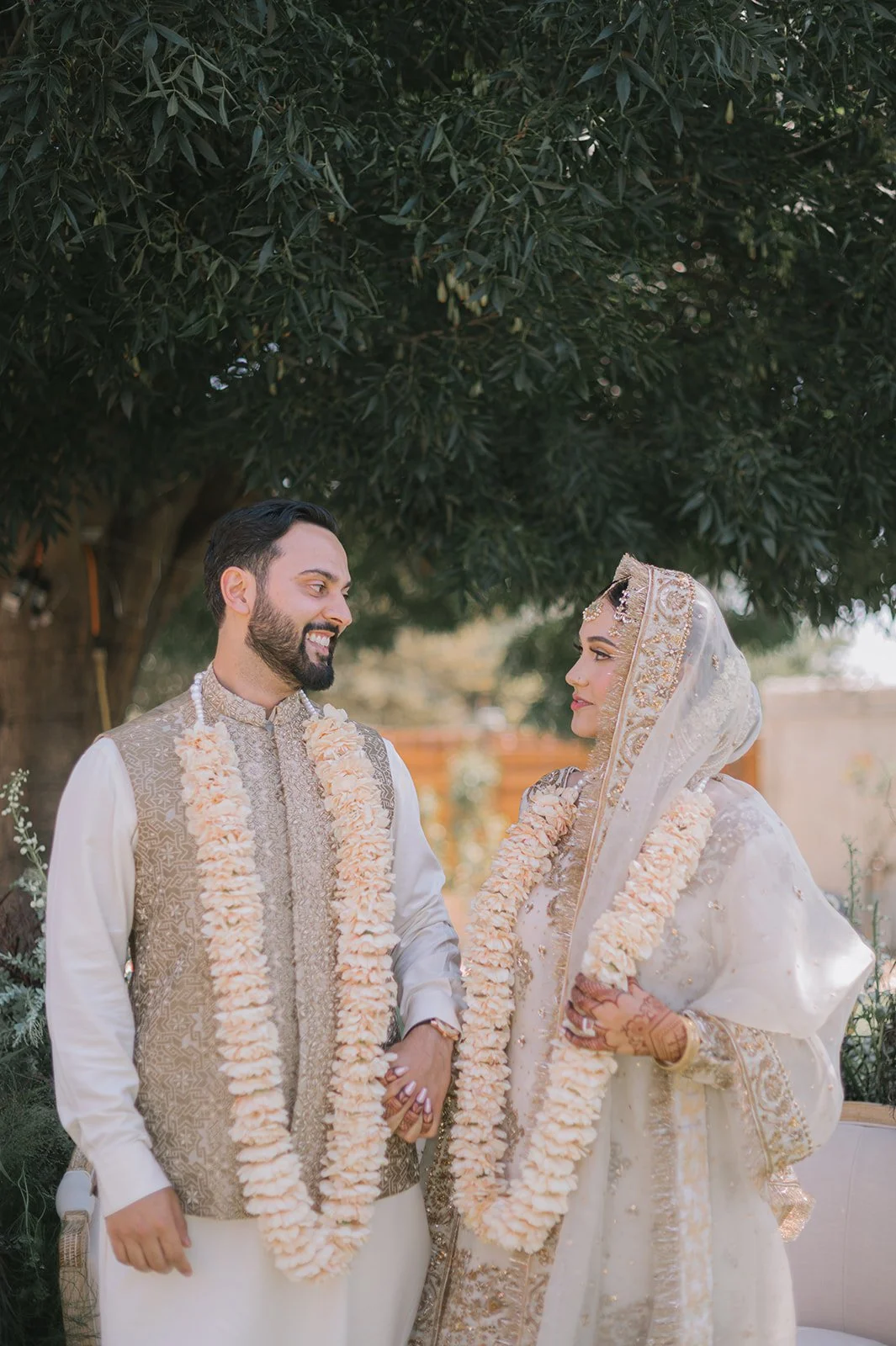 A bride and groom in traditional wedding attire, wearing flower garlands, standing outdoors under a tree, looking at each other.