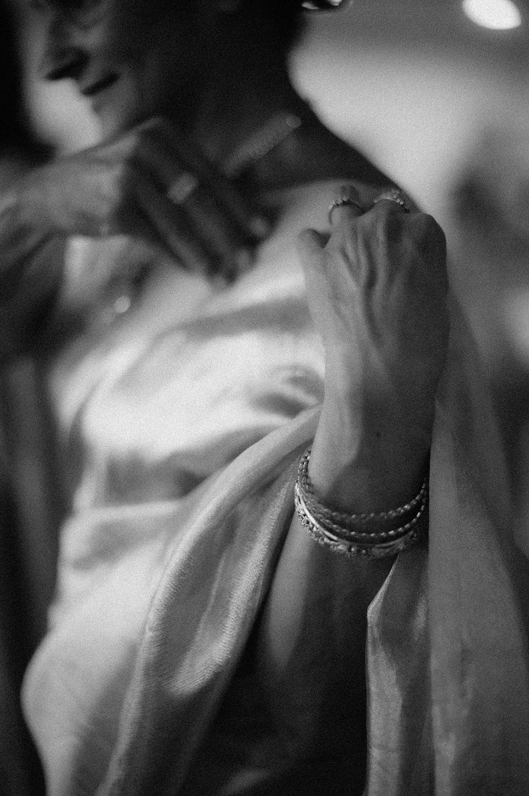 Close-up black and white photo of a woman wearing jewelry, with a visible hand and wrist adorned with bracelets, as she drapes a fabric over her shoulder.