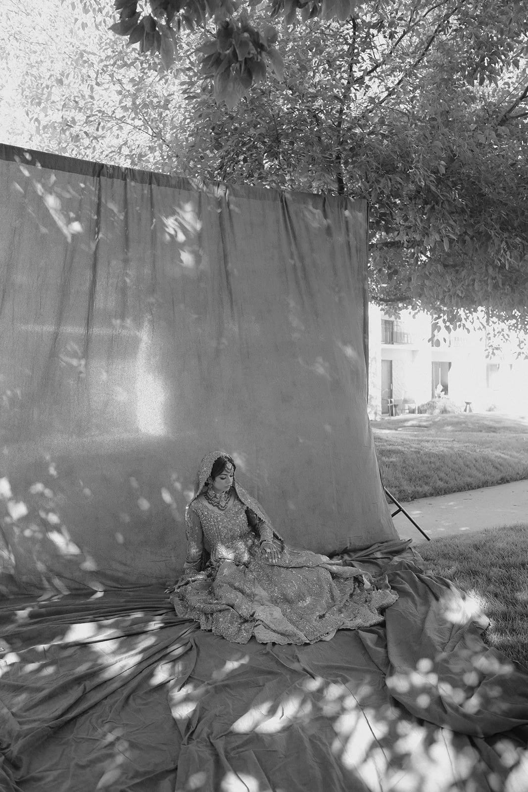 A woman in traditional bridal attire sitting on fabric in front of a large backdrop outdoors, with trees and buildings visible in the background.