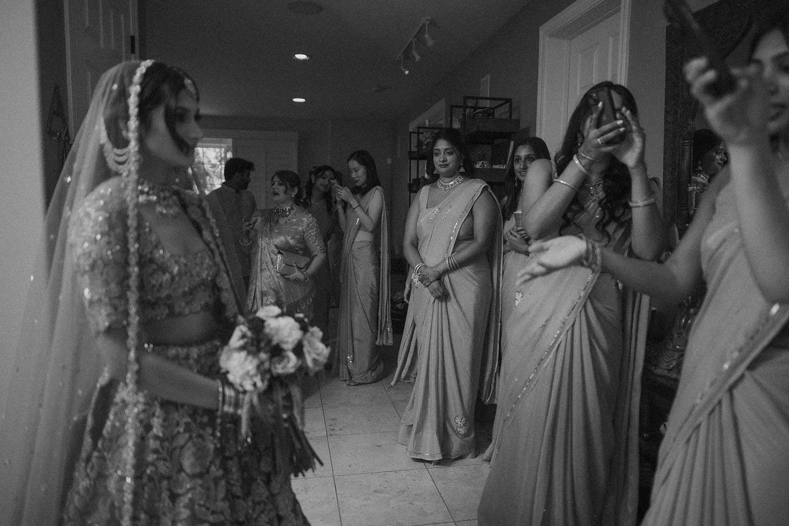 A bride in traditional Indian attire holding a bouquet, surrounded by women in sarees at a wedding ceremony.