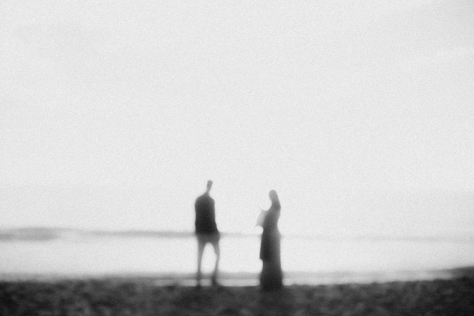 Two people standing on a beach, facing each other, blurred and in black and white.