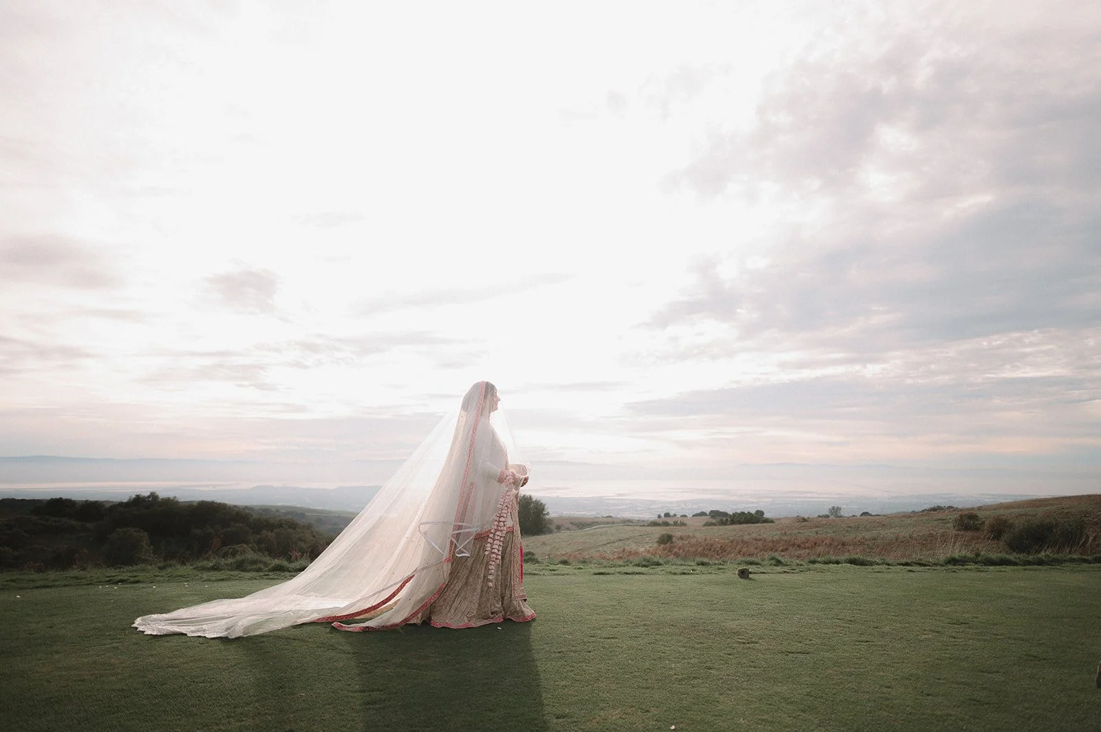 A bride in a wedding dress and veil standing outdoors on a grassy hill, overlooking a distant landscape under a cloudy sky.