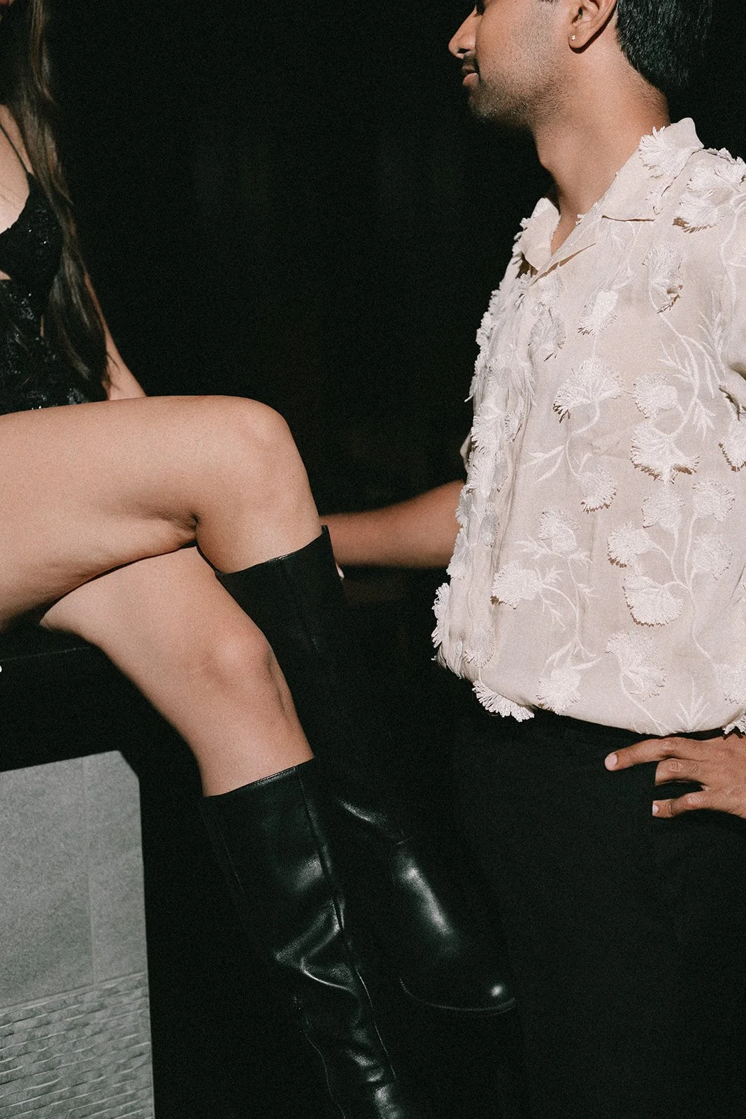 A man and woman are engaged in an intimate moment, with the woman sitting on a surface and the man facing her. The woman is wearing a black outfit and black knee-high boots, while the man is dressed in a white embroidered shirt and black pants.