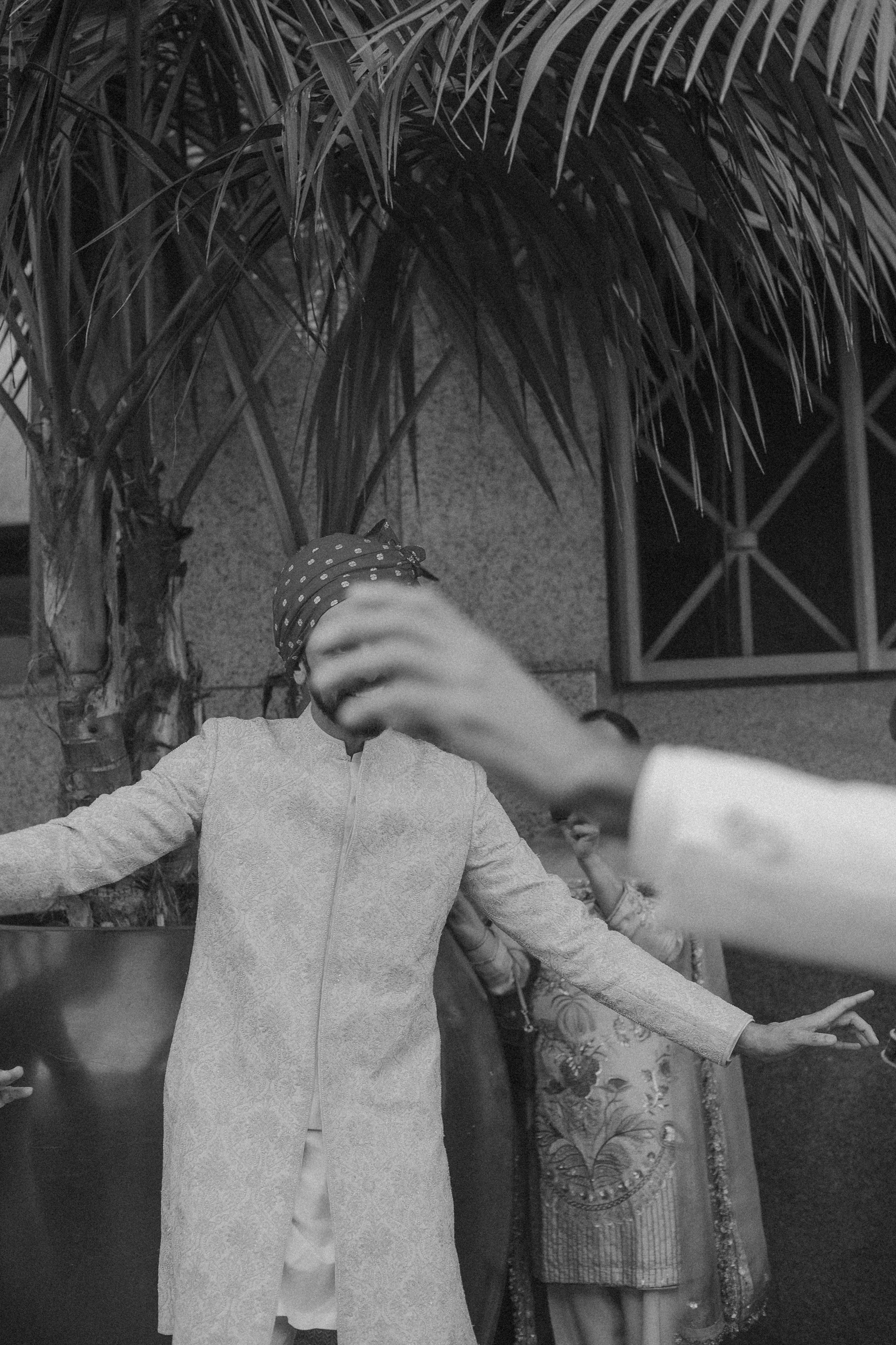 Black and white photo of a person wearing a long coat and polka dot headscarf, with their arms outstretched and their face obscured by their hand, standing in front of a tree and a building with a window.