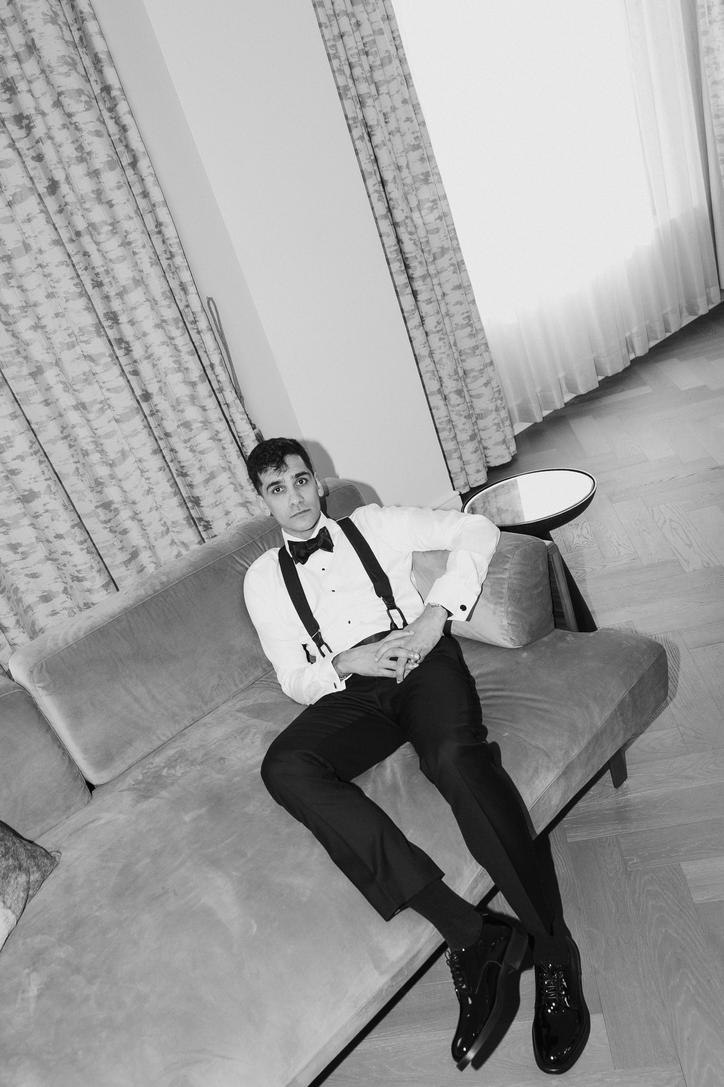 A man sitting on a sofa, dressed in formal attire with a bow tie, suspenders, and shiny black shoes, in a room with curtains and a small side table.