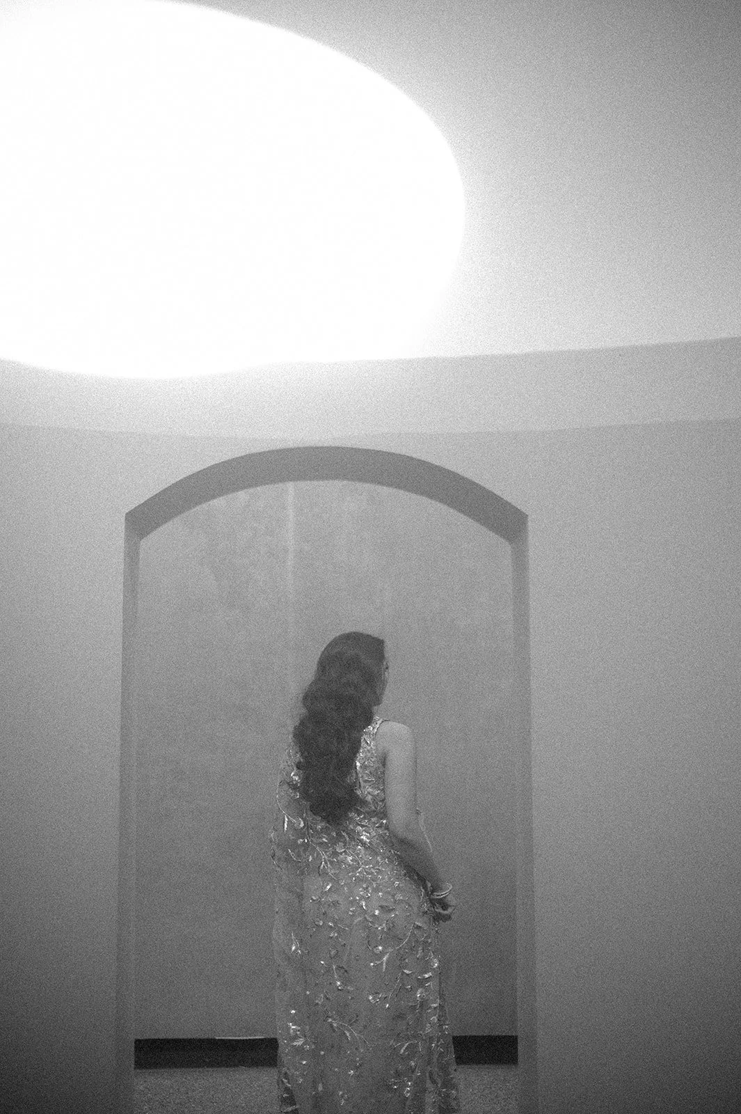 A woman with long, wavy hair wearing a sleeveless, embellished gown, standing with her back to the camera in front of an arched wall, illuminated by a bright ceiling light.