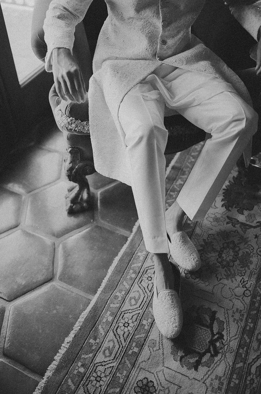 A person wearing traditional South Asian attire, sitting on an ornate chair with legs crossed. The person is dressed in light-colored, textured fabric, with a long tunic and loose pants. They are wearing footwear that looks textured, and their hand is resting on the armrest of the chair.