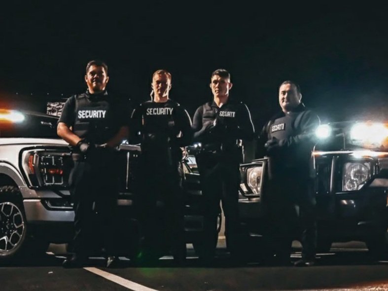 Four security guards standing in front of SUVs at night, wearing black uniforms with 'Security' written on them.