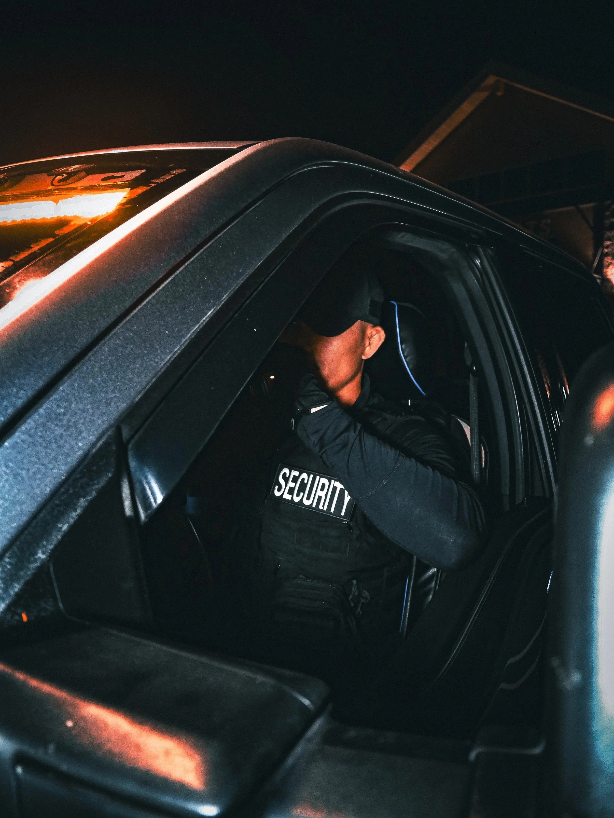 A security guard sitting inside a dark vehicle at night, wearing a black uniform with the word 'SECURITY' on the vest, and adjusting a cap while looking down.