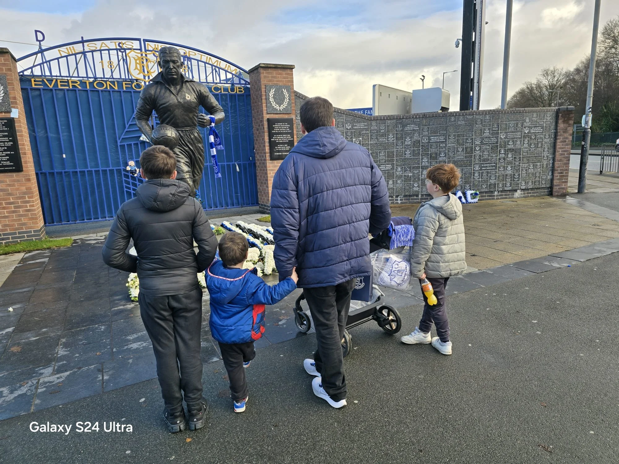 People gathered around a memorial with a bronze statue of a football player at Everton Football Club, with banners, flowers, and inscriptions on the memorial wall behind.