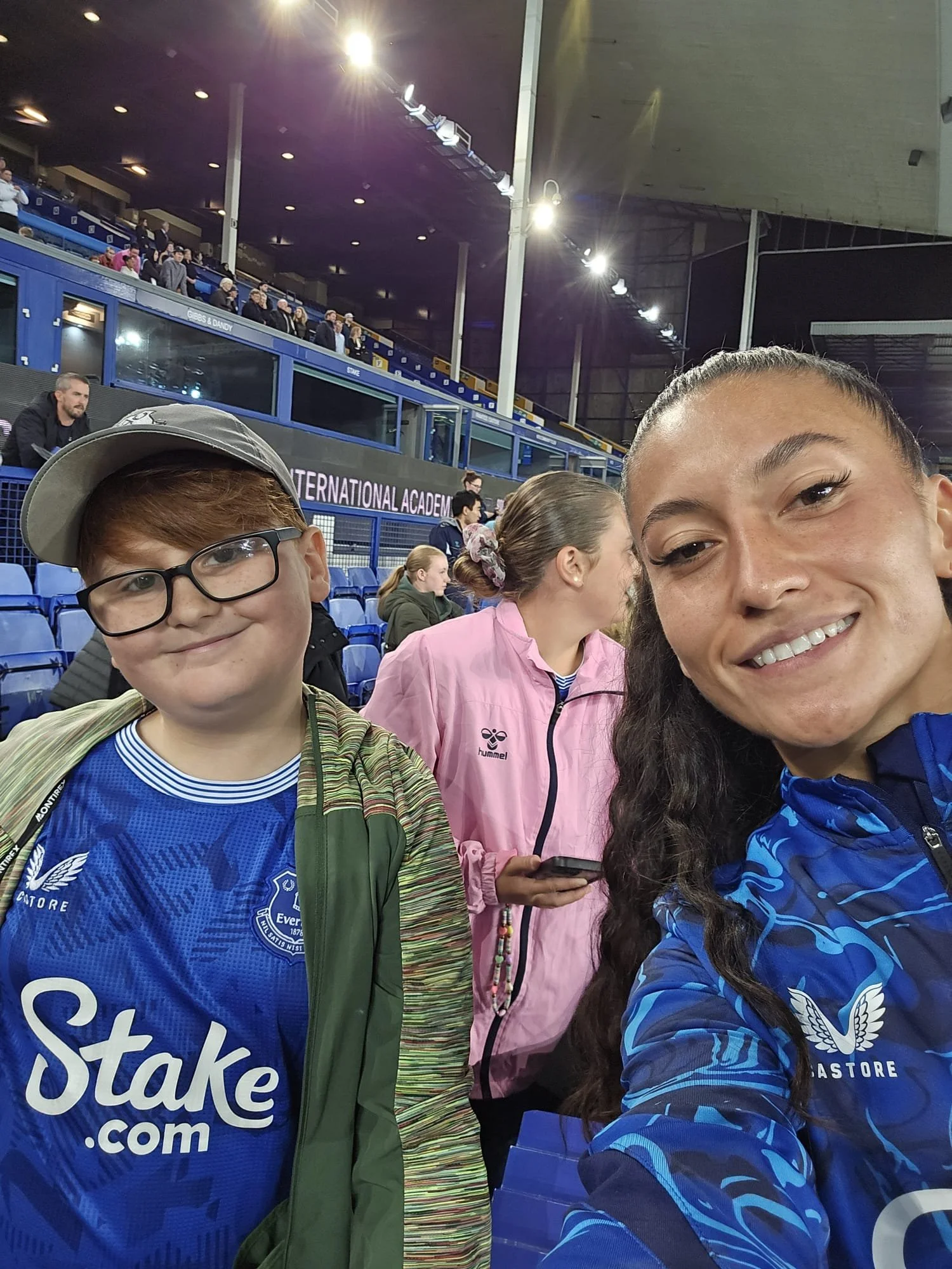 Two women and two children at a sports event in a stadium. The woman on the right is smiling and wearing a blue sports jacket, the woman in the middle in pink, the boy on the left is wearing glasses and a sports jersey, and the girl in the middle has a bun and is using a phone. The stadium seating and spectators are in the background.