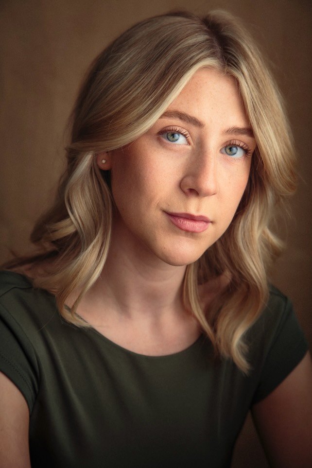 A young woman with blonde hair styled in loose waves, blue eyes, and fair skin, wearing a dark green top and small earrings, looking at the camera against a brown background.