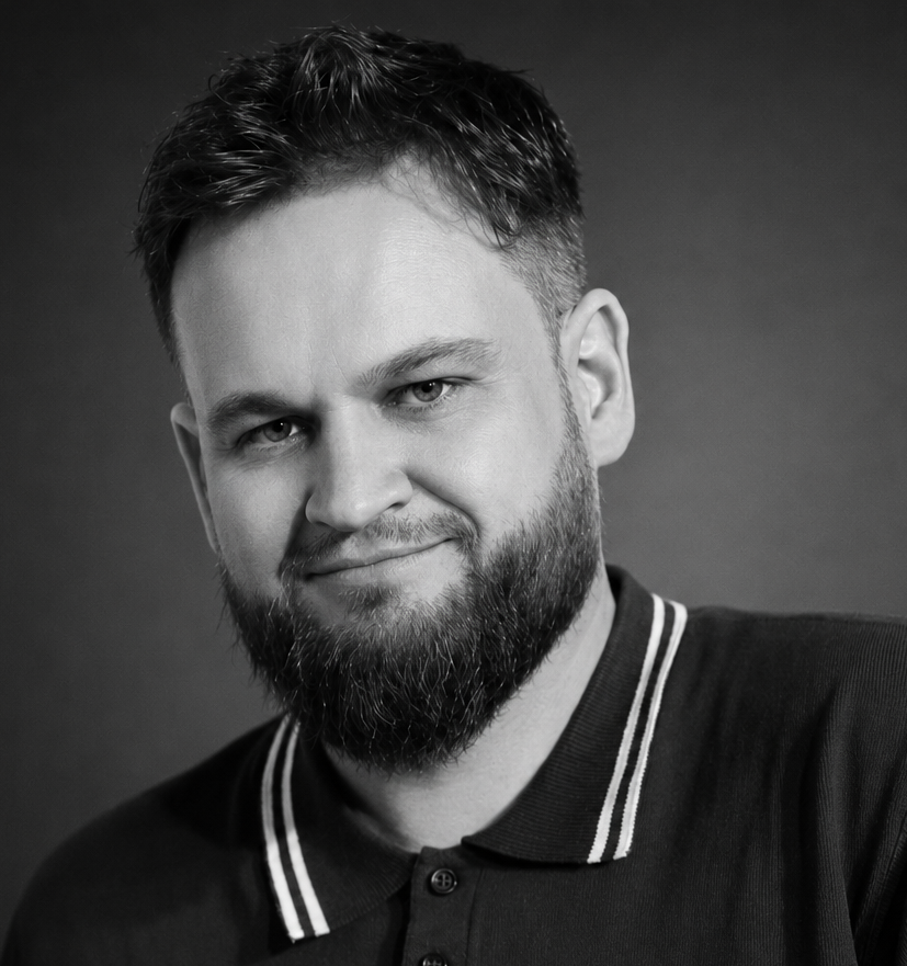 Black and white portrait of Adam Hardaker with short hair, a beard, and a slight smile, wearing a collared shirt with striped trim.