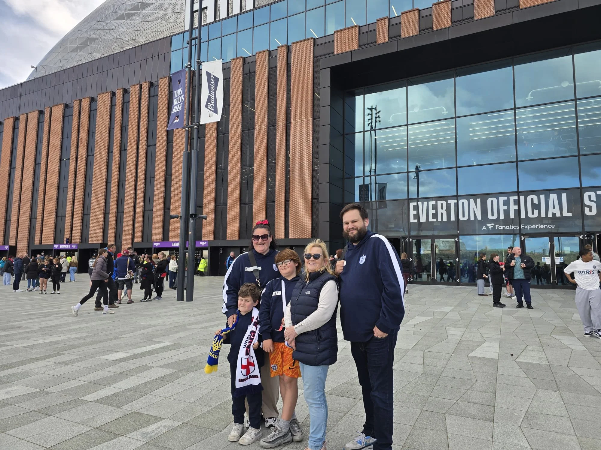 Group of fans standing outside Everton Stadium, some wearing England soccer merchandise, with others in casual clothing, and the stadium's modern exterior with large glass windows and brick accents in the background.