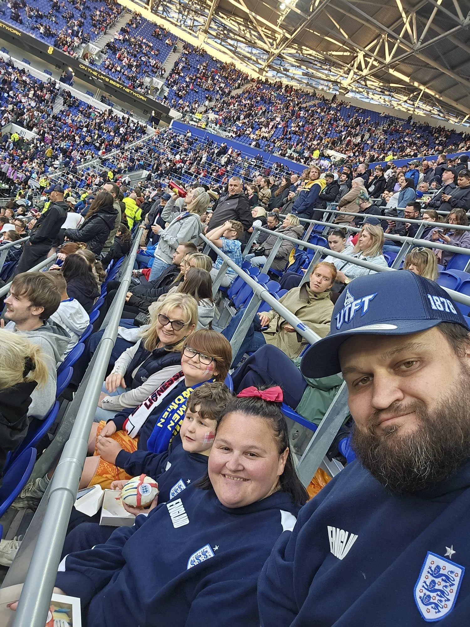 Crowd of people seated in a stadium, some wearing England football team jerseys, in a large indoor arena with blue seats and a high ceiling.