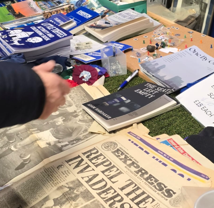 A cluttered table with pamphlets, documents, a book titled 'The Seattle Left Empty,' a newspaper headline 'Reflections Reported.' There are small pins, a pen, and various papers. A person's hand is partially visible reaching towards the items.