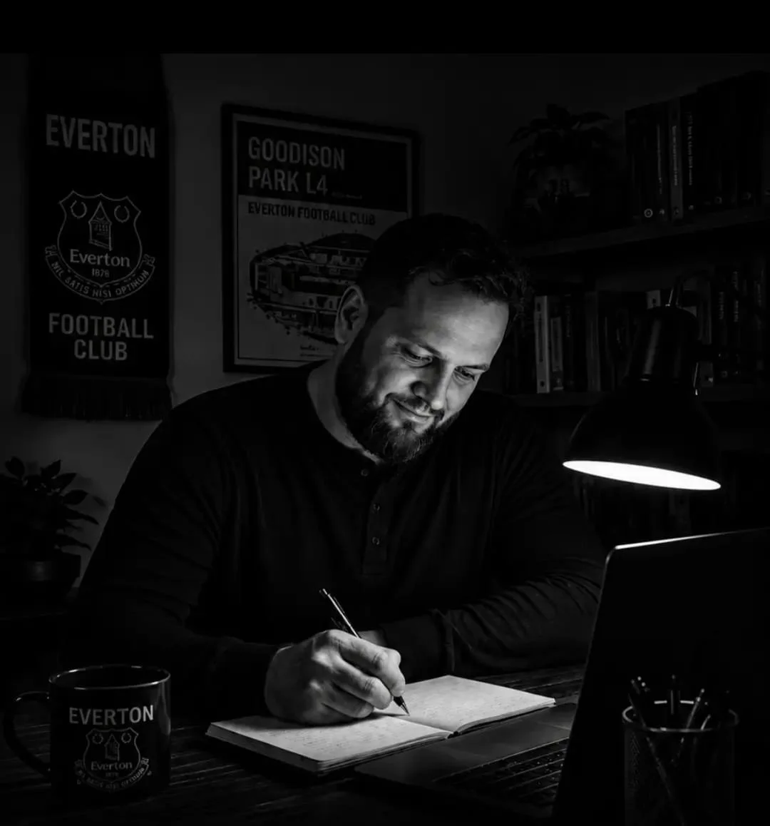A man working at a desk in a dimly lit room, writing in a notebook with a laptop in front of him, and an Everton Football Club mug nearby. Bookshelves and posters are visible in the background.