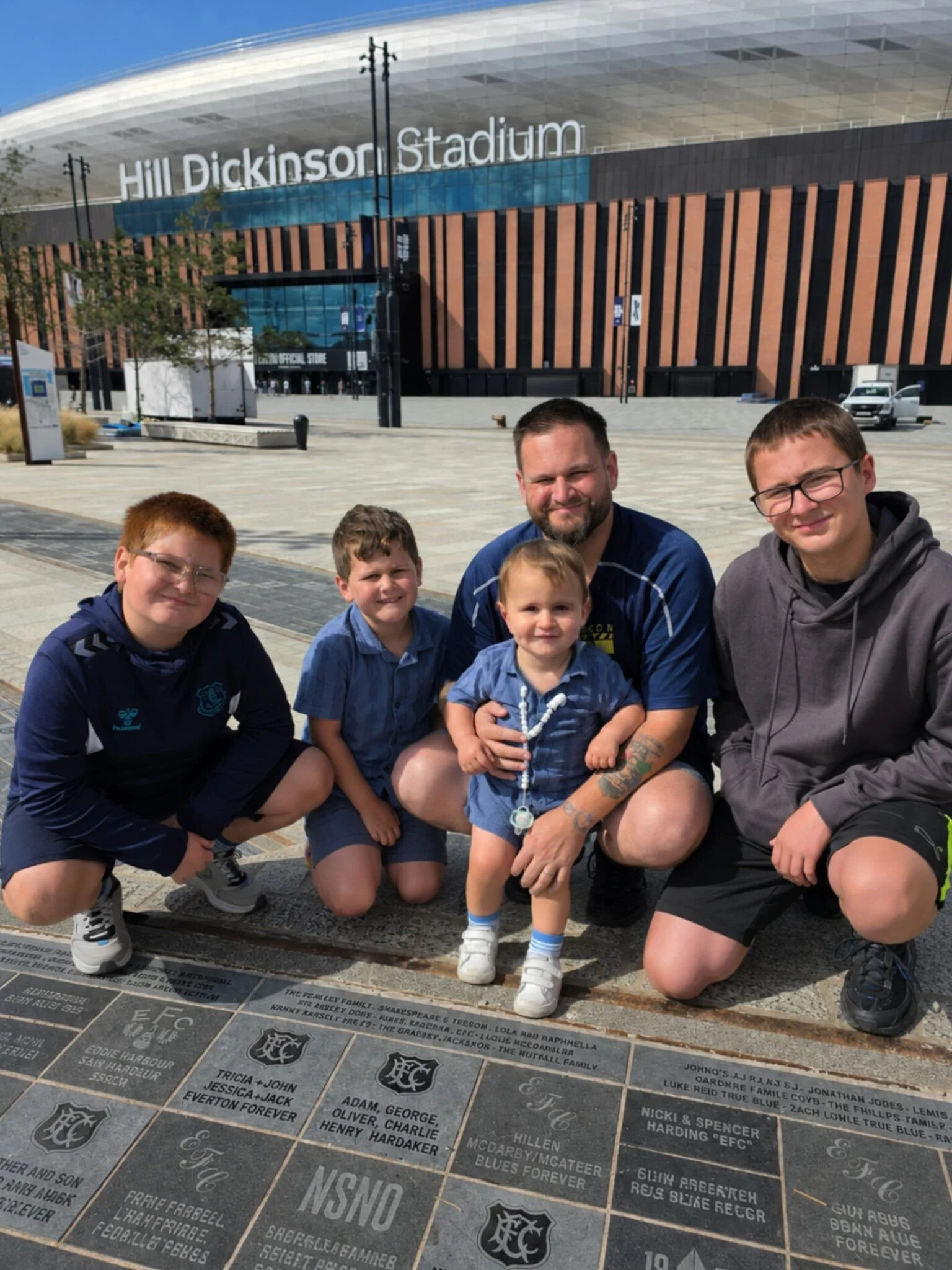 A family of five posing together in front of the memorial at Hill Dickinson Stadium, with the stadium in the background.