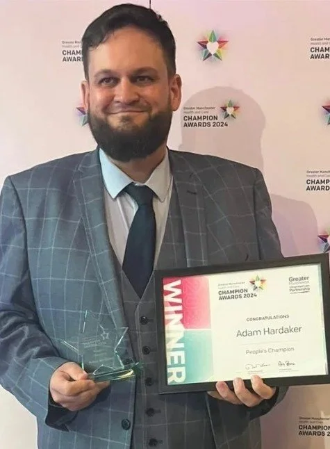 Adam Hardaker in a gray checkered suit and blue tie holding a framed award certificate that says 'Winner' and 'Adam Hardaker People's Champion' at the 2024 Champion Awards event, with a pink and white background featuring the event's logo and text.