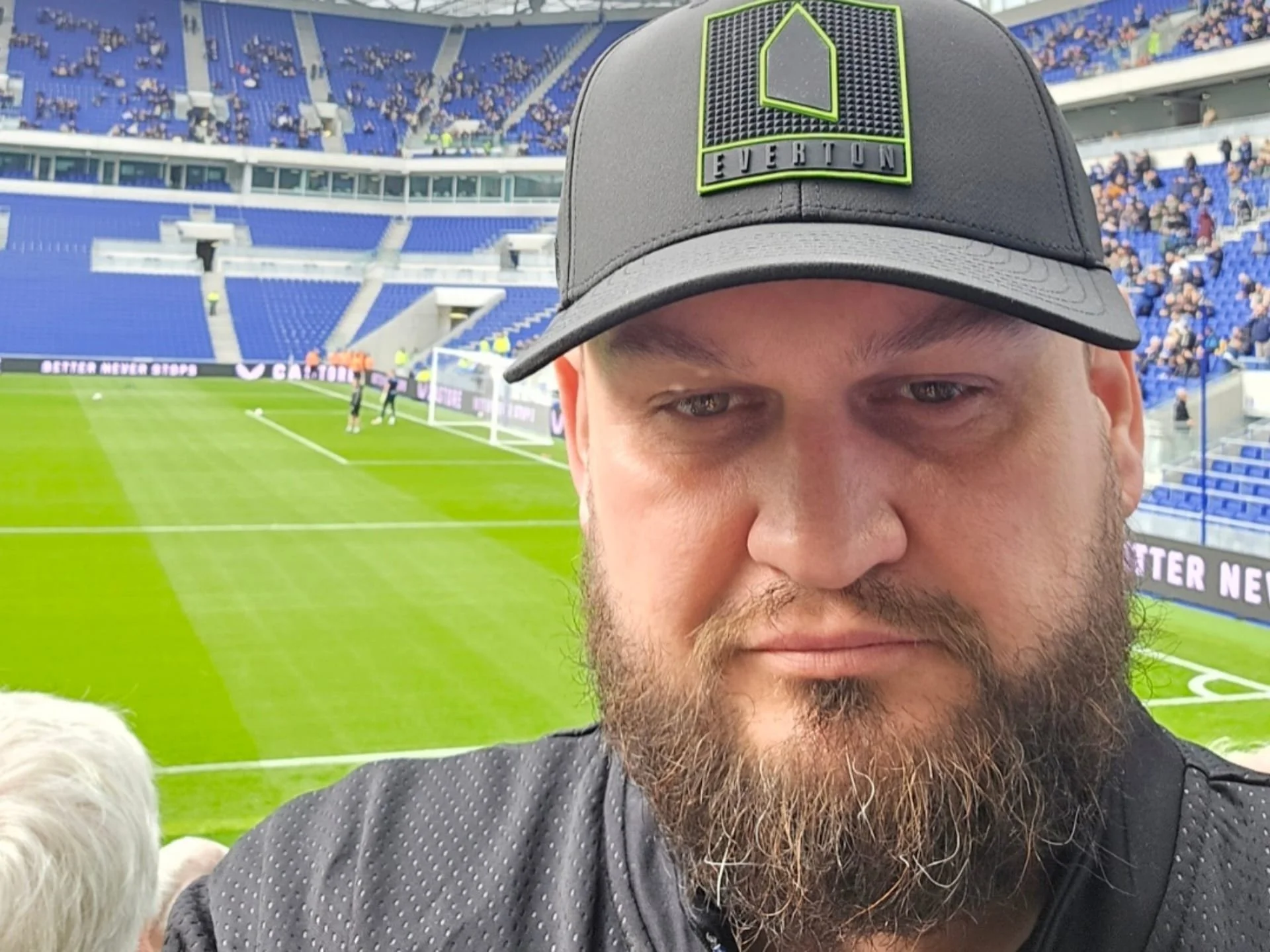 A man with a beard wearing a black cap with a green and black logo, taking a selfie at a soccer stadium with blue seating and a green field in the background.