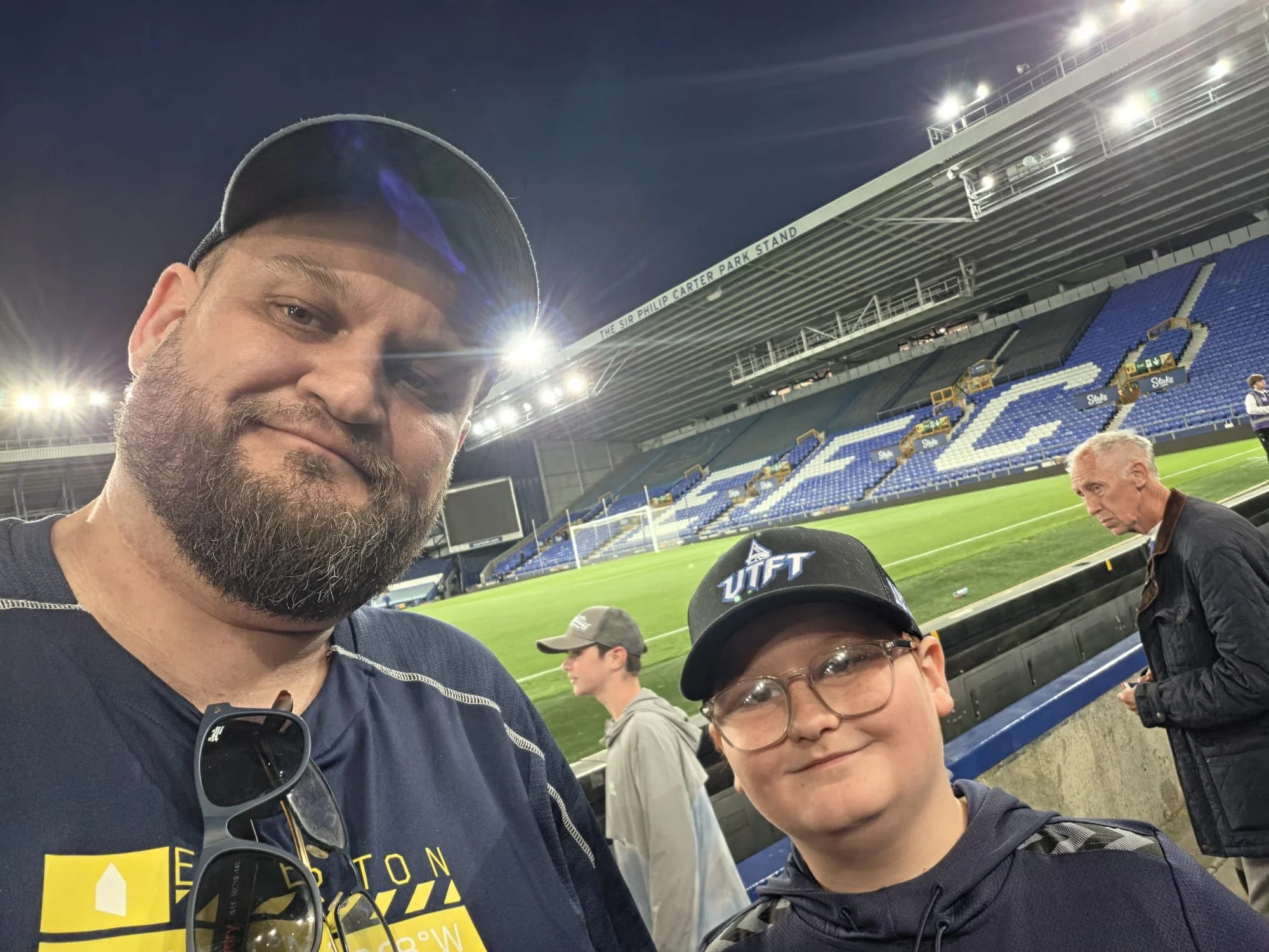 Two men taking a selfie at a stadium during nighttime, with empty blue and white seats in the background. The man in the foreground has a beard, wears a cap and sunglasses hanging from his shirt, and is smiling slightly. The second person, partly visible, wears glasses and a jacket. The stadium lights are bright, and the words "The Sir Philip Carter Park Stand" are visible on the upper part of the stands.