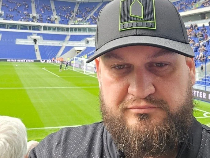 Man with a beard taking a selfie at a soccer stadium with a green field and players in the background.