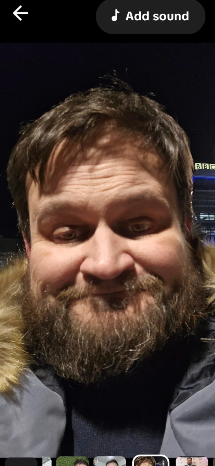 Close-up of a bearded man with brown hair at night, outdoors, with a dark sky and a building with the BBC logo in the background.