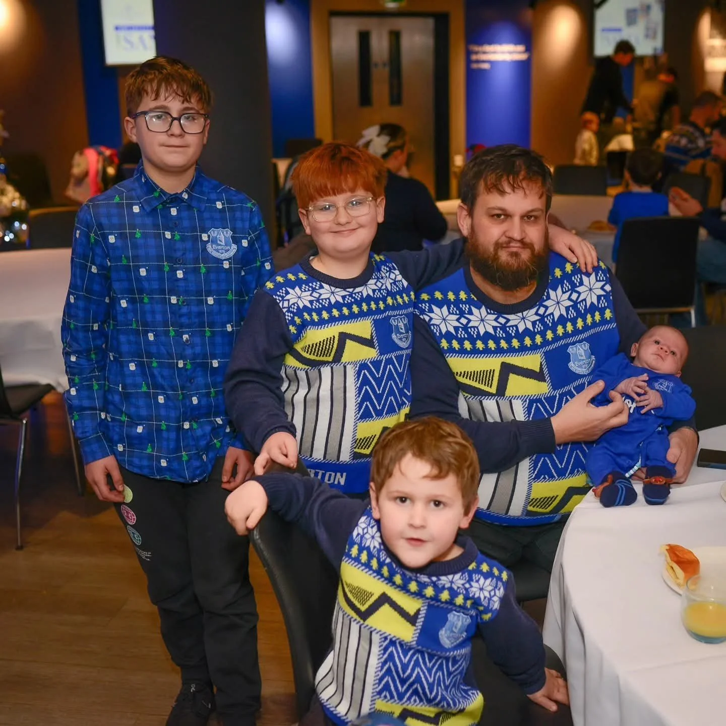 A group of five people, including three children and two adults, gathered around a table at a festive event, with Christmas decorations and other people in the background. They are all wearing holiday-themed clothing, with some wearing sweaters and shirts displaying a sports team's logo.