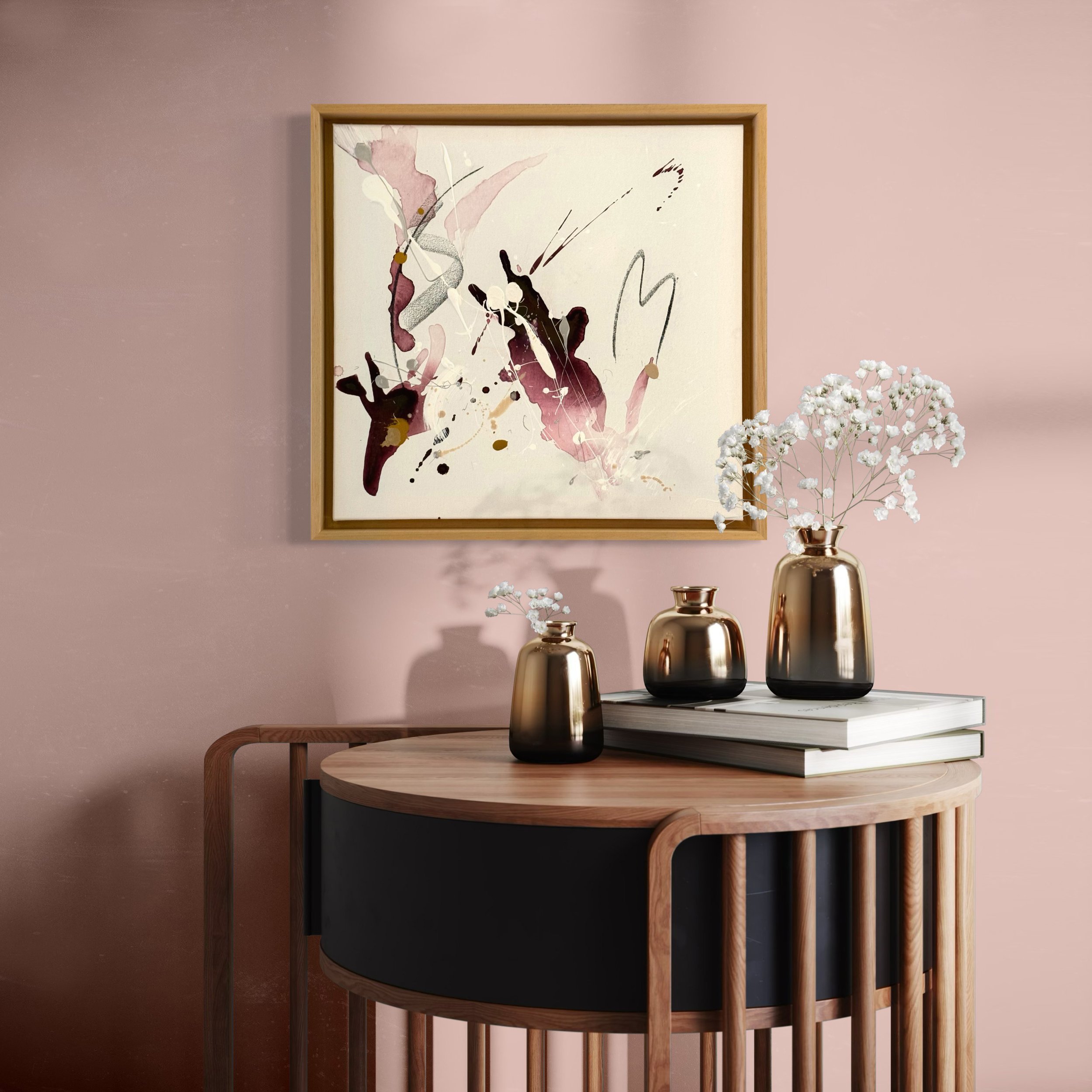 Ein Wandbild mit abstrakten Pinselstrichen in Rosa, Schwarz, Grau und Gold hängt an einer rosa Wand. Darunter stehen drei Vasen in Bronze-Optik, mit weißen Blumen, auf schwarzen und weißen Büchern auf einem runden Holztisch.