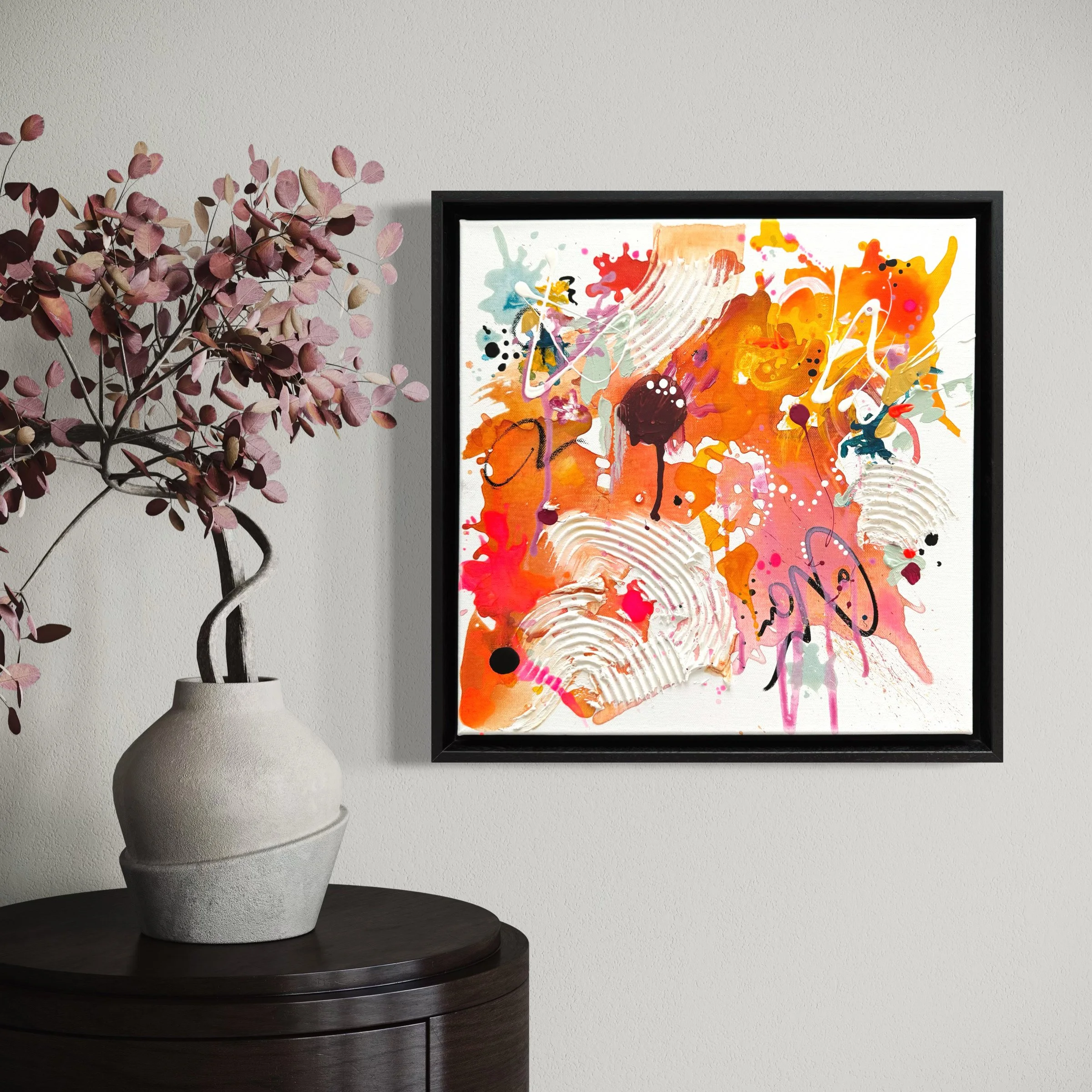Moderne abstrakte Malerei mit bunten Farbklecksen und Pinselstrichen, in schwarzen Rahmen gehalten, an einer weißen Wand. Neben einer Vase mit Pinkfarbenem Blätterzweig auf einem dunklen Holz-Tisch.