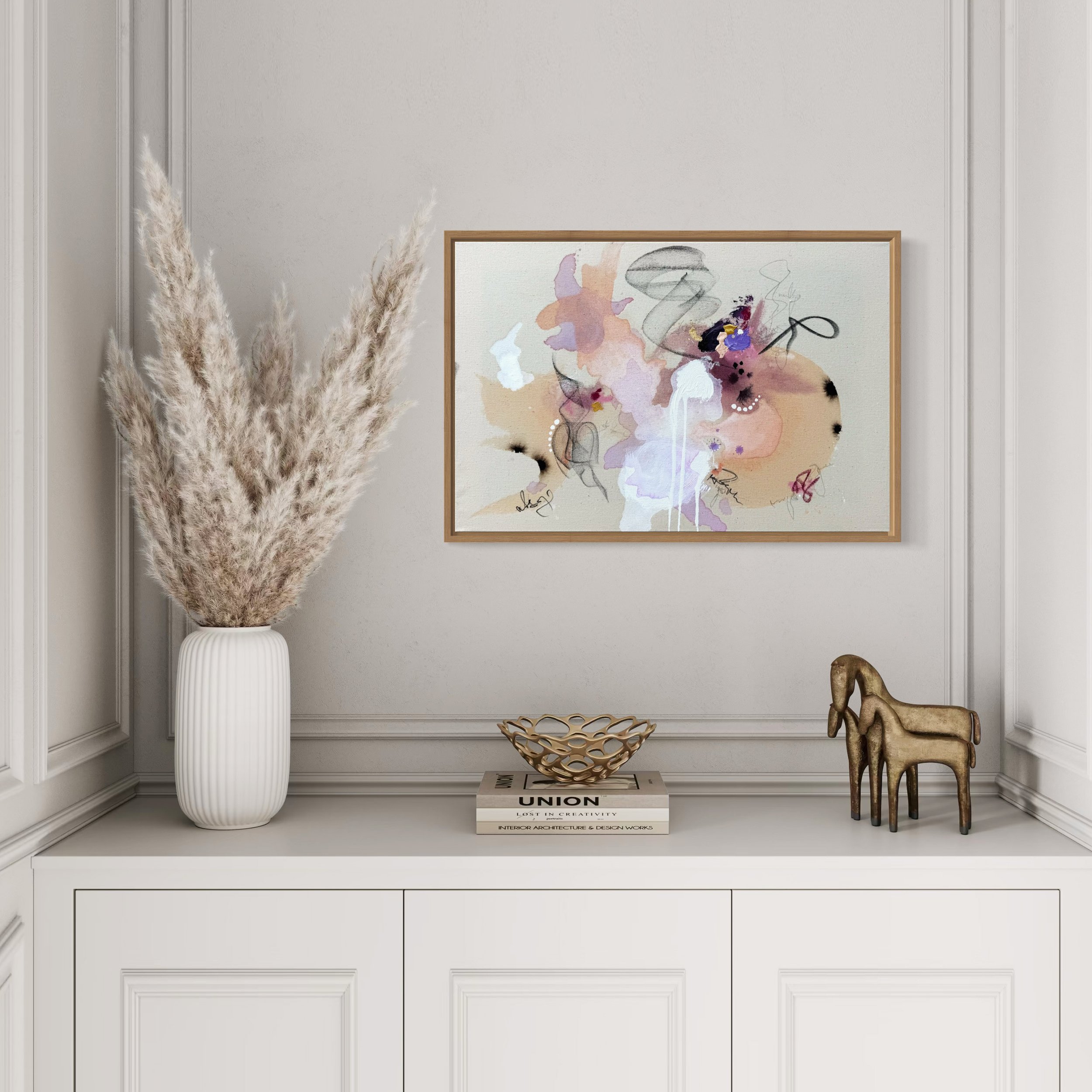 Dekoratives Sideboard mit einer Vase voller Pampasgras, einem Buch, einer goldenen Skulptur und einer kleinen Metallfigur eines Pferdes, über einem abstrakten Gemälde in einem weißen Raum.