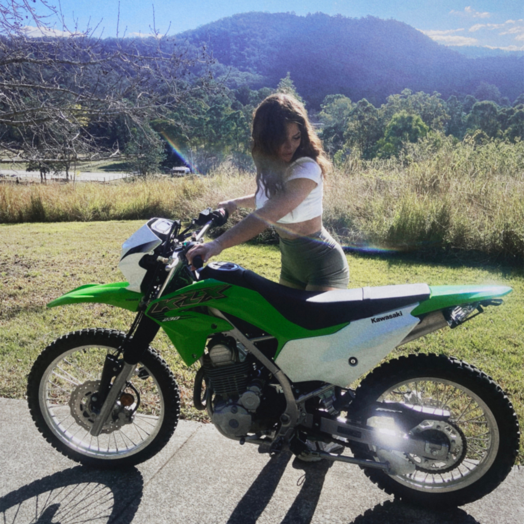 A woman with long hair, wearing a white crop top and shorts, standing beside a green Kawasaki dirt bike on a sunny day in a grassy area with trees and mountains in the background.