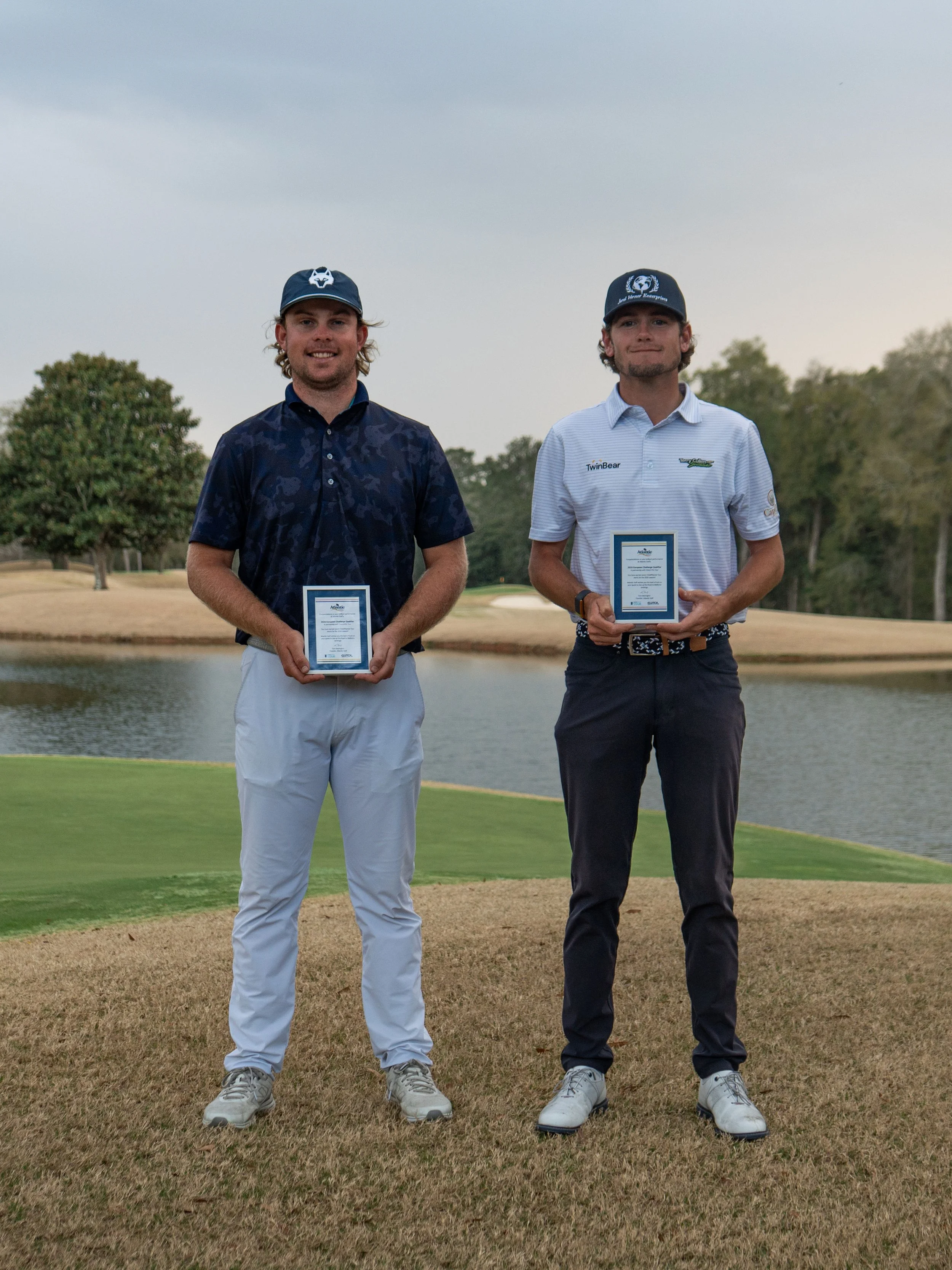 A golfer in white shirt, dark shorts, hat, and sunglasses preparing to swing on a golf course with rolling green hills, mountains, and cloudy sky in the background. There are advertising signs for hotelplanner TOUR and Road to Mallorca.