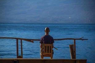 A person sitting on a chair by the water, facing the ocean with a fishing rod, during daytime.