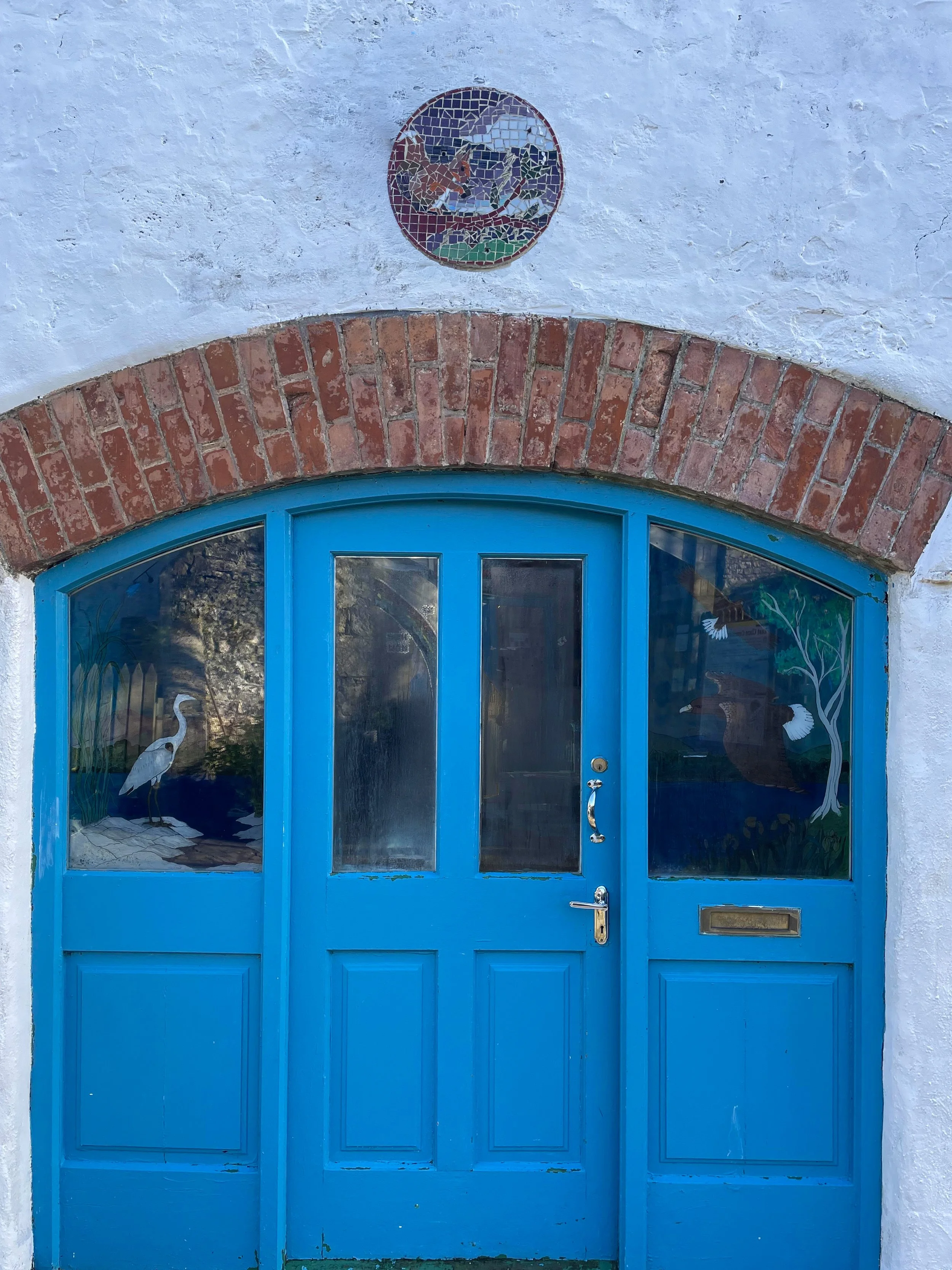 Blue door with a glass window, set in a white stucco wall with a red brick arch above. The glass features a painted scene of a heron, a tree, and a flowering plant.