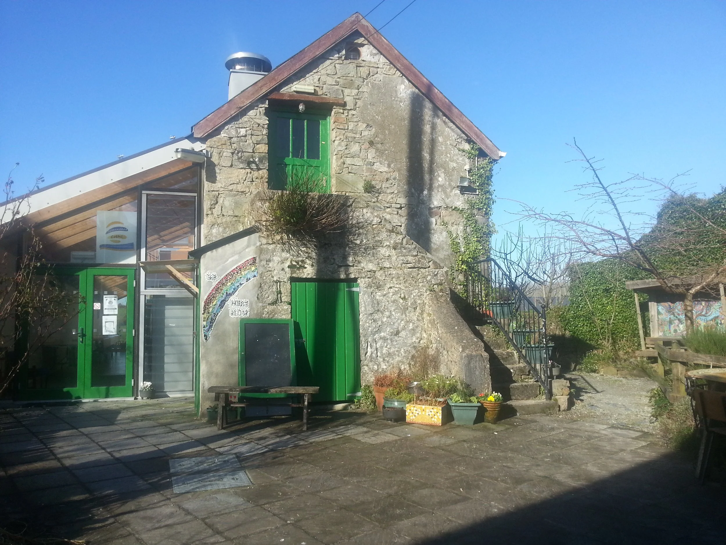 A rustic stone house with a steep roof, green doors and windows, surrounded by potted plants and outdoor furniture, under a clear blue sky.