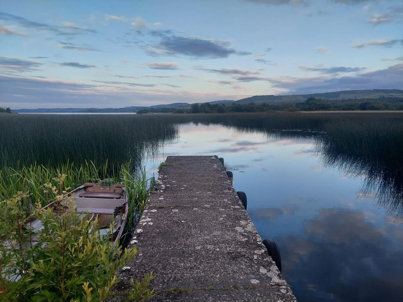 A narrow concrete pier extends into a calm lake with tall grasses on each side. The sky has scattered clouds, reflecting in the water with distant rolling hills in the background.